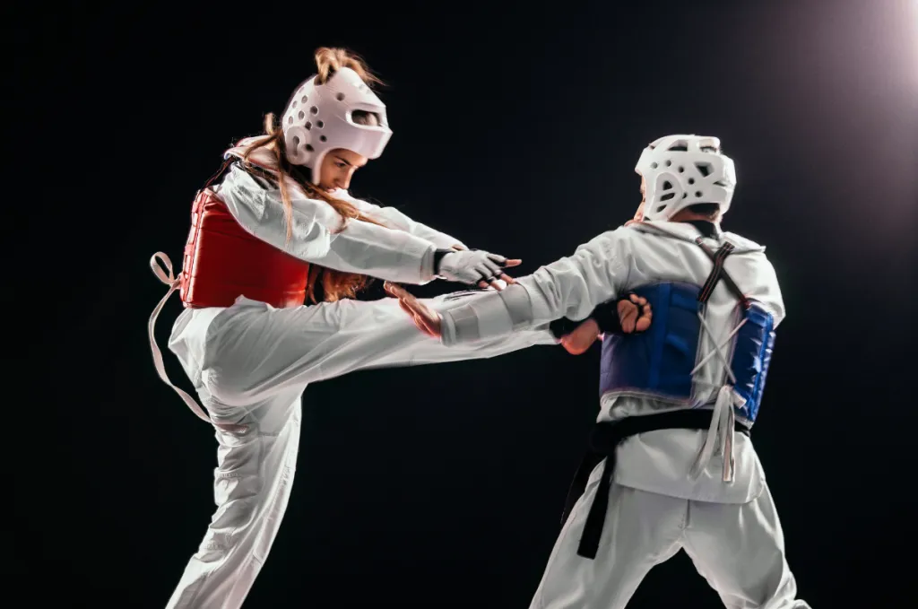 Two taekwondo athletes in headgear and chest protectors sparring, one throwing a high kick