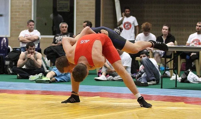 Two wrestlers grappling on a mat in a training hall while others watch from the benches