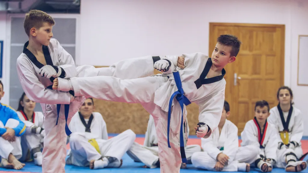 Boy in a taekwondo dobok holding a high kick against an instructor's pad while classmates watch
