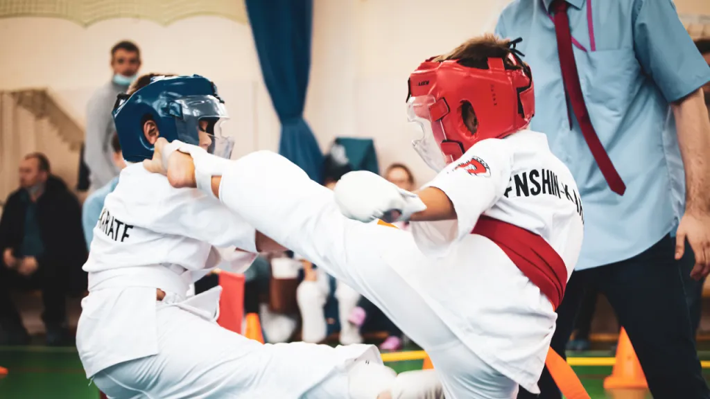 Two young martial artists in headgear and gloves sparring at a tournament while the referee watches